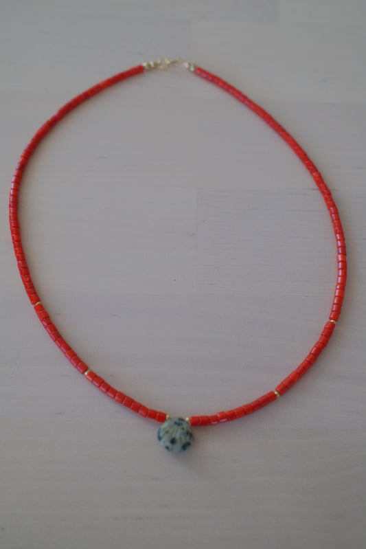 A red beaded necklace with a small white and blue patterned bead accent and tiny gold spacer beads, displayed on a light off-white background.