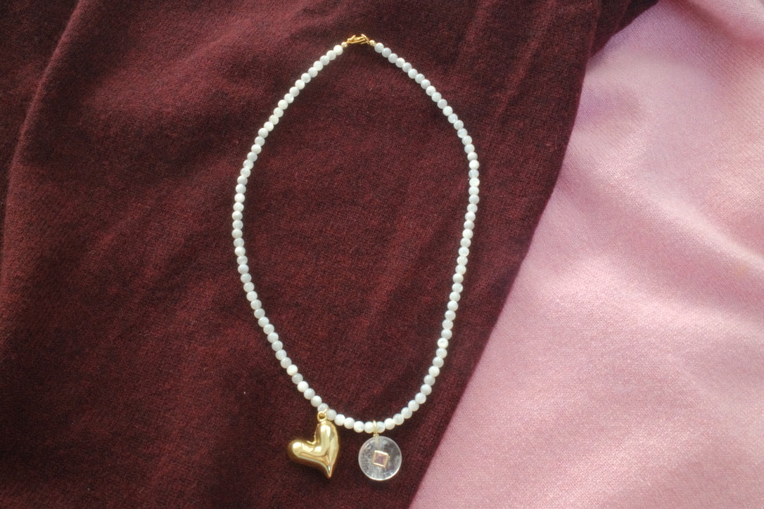 Pearl necklace with a gold heart charm and silver round charm on a textured surface.