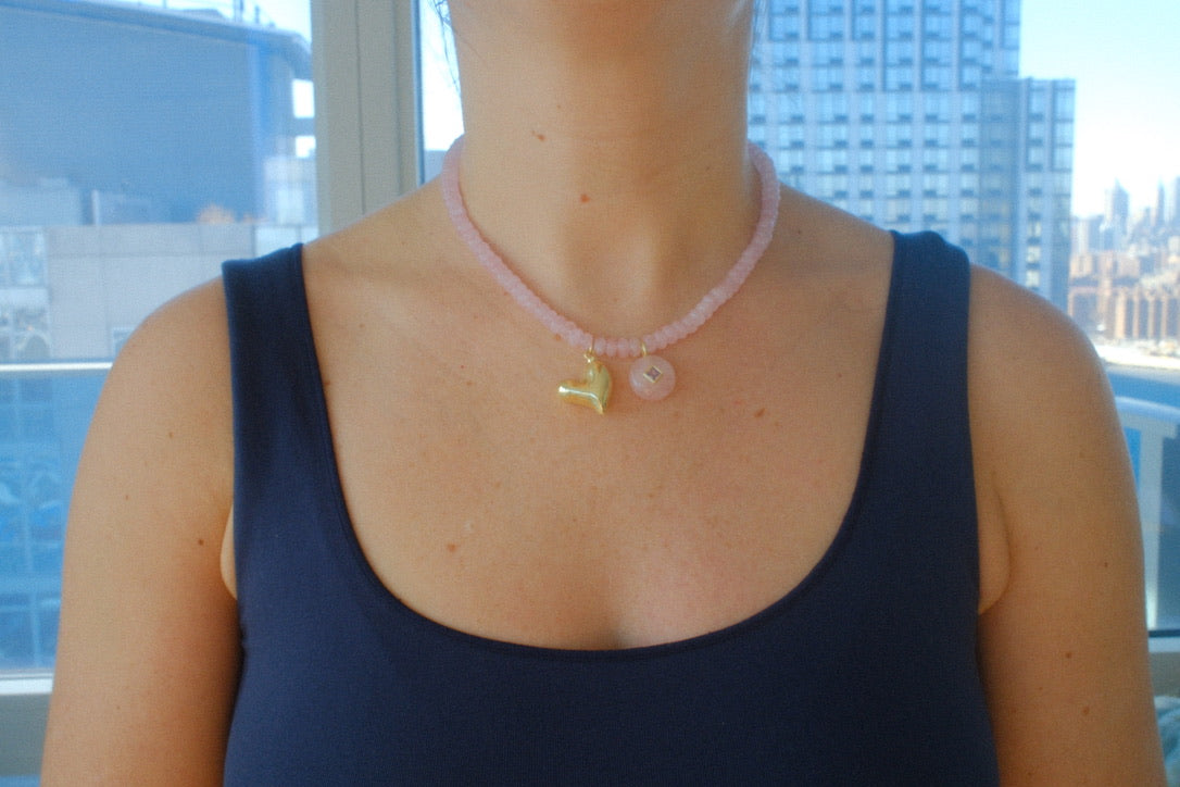 Person wearing a pink beaded necklace with heart charm in front of a cityscape