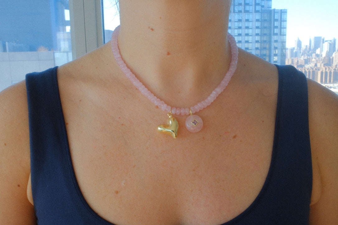 Necklace with a gold heart pendant and pink beads worn by a person in front of a cityscape.