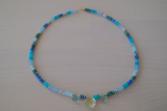 A beaded necklace with small turquoise, blue, green, and white beads, featuring a central faceted pale yellow crystal pendant, displayed on a light beige wooden surface.