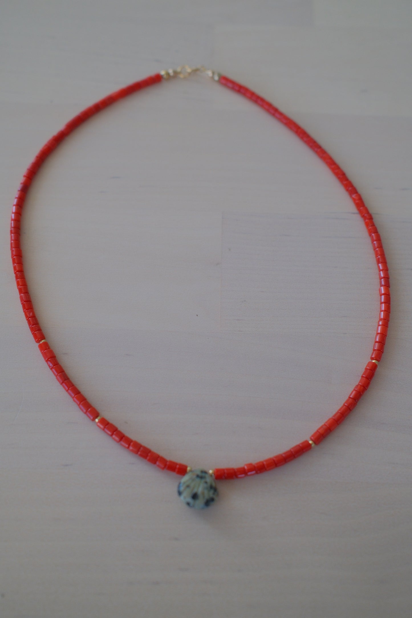 A red beaded necklace with a small white and blue patterned bead accent and tiny gold spacer beads, displayed on a light off-white background.