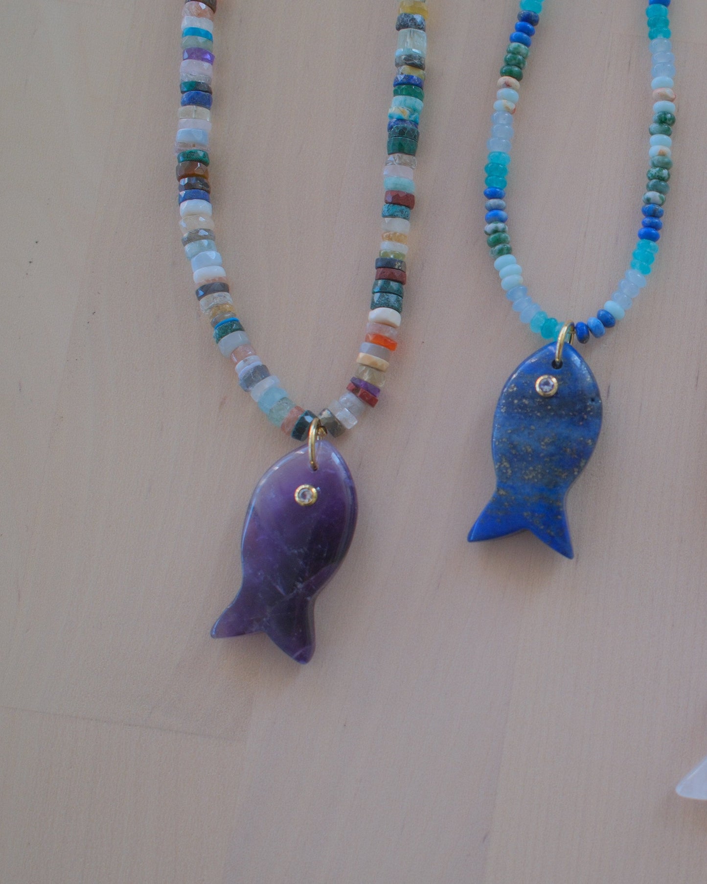 Four beaded fish necklaces with colorful seed beads and fish pendants in purple, blue, white, and green, displayed on a light beige background.