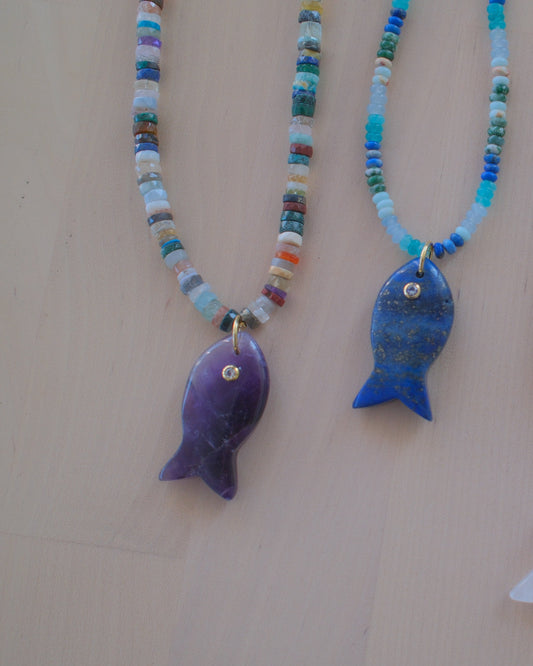 Four beaded fish necklaces with colorful seed beads and fish pendants in purple, blue, white, and green, displayed on a light beige background.
