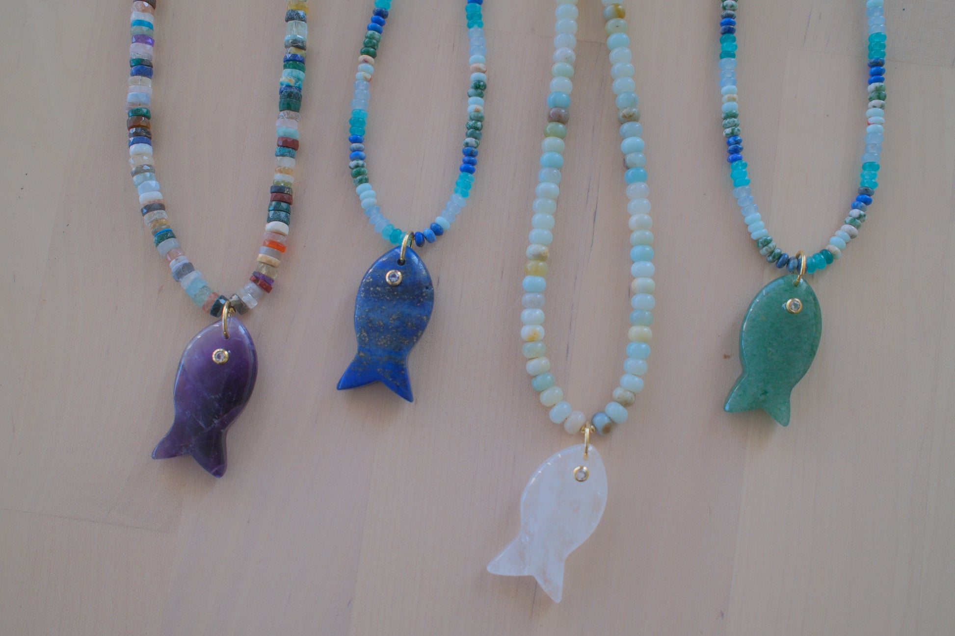 Four beaded fish necklaces with colorful seed beads and fish pendants in purple, blue, white, and green, displayed on a light beige background.