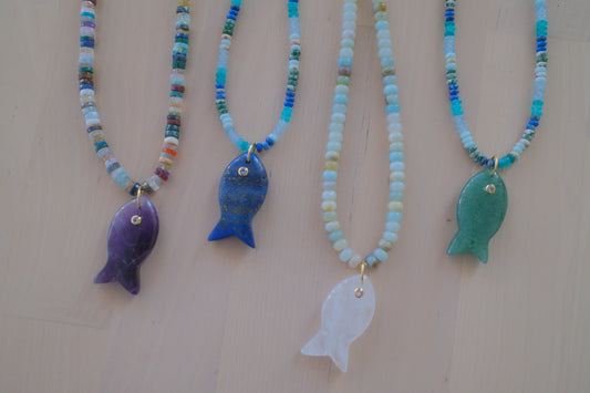 Four beaded fish necklaces with colorful seed beads and fish pendants in purple, blue, white, and green, displayed on a light beige background.