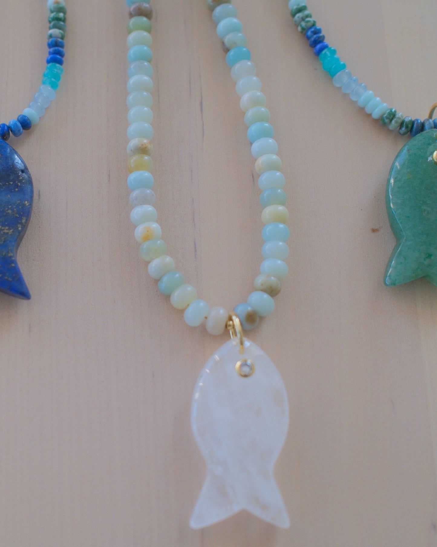 Four beaded fish necklaces with different stone fish pendants (purple, blue, white, and green) displayed on a light wood background.