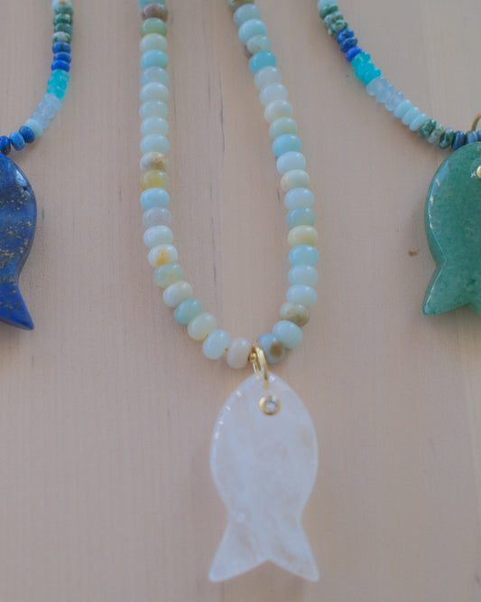 Four beaded fish necklaces with different stone fish pendants (purple, blue, white, and green) displayed on a light wood background.
