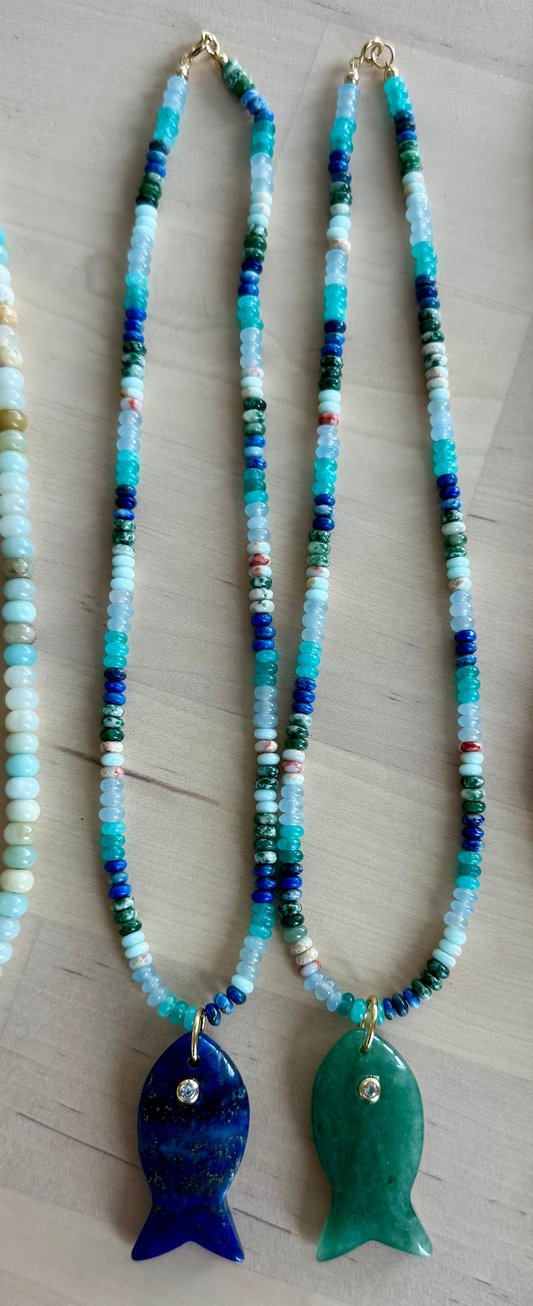 Four beaded fish necklaces arranged side by side on a light wood surface. The necklaces feature different bead patterns and fish pendants: a white shell pendant, a dark blue pendant, a green pendant, and a purple pendant.